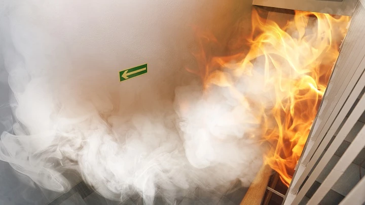 Lågor från brand och rök syns i ena delen av rummet. På väggen syns utrymningsskylt.